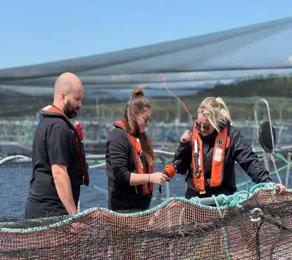Two women in orange life jackets work with fishing nets on a boat, with aquaculture pens visible in the background.