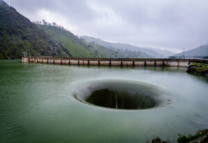 Large circular spillway creating a dramatic whirlpool in a reservoir, with a concrete dam and green hills in the background.