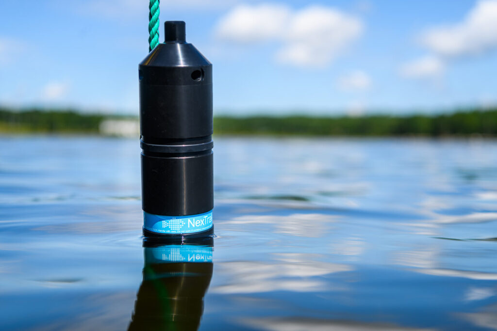 A black cylindrical water quality monitoring device with blue "NexTrak" branding floats partially submerged in calm blue water under a cloudy sky.