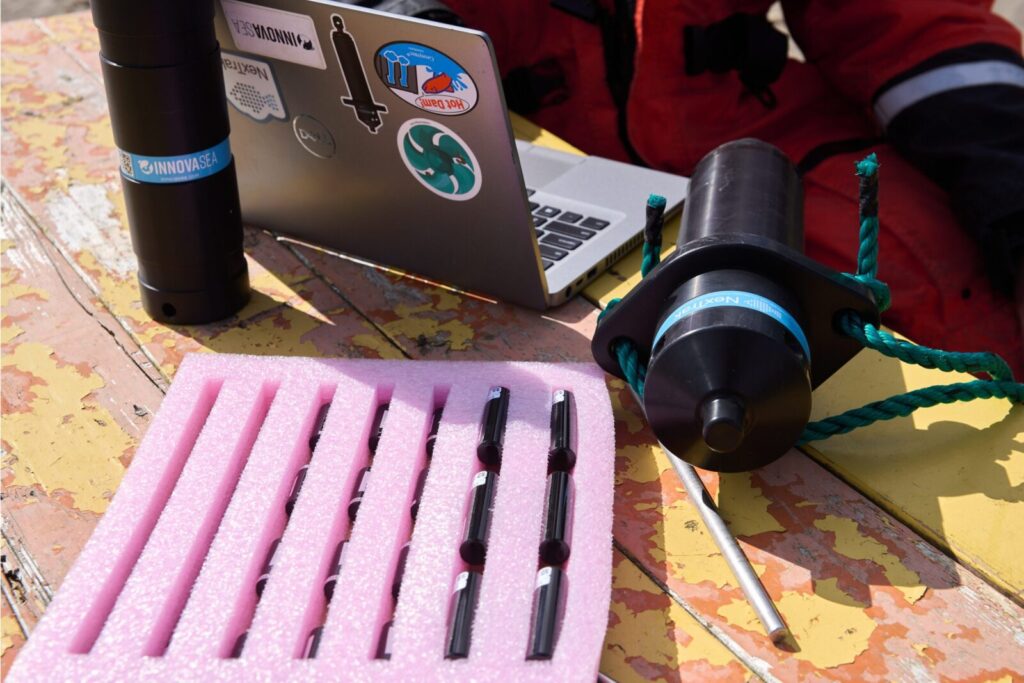 Scientific equipment on weathered wooden deck including laptop with stickers, black cylindrical device with rope, and pink foam holder with dark rod-like instruments.