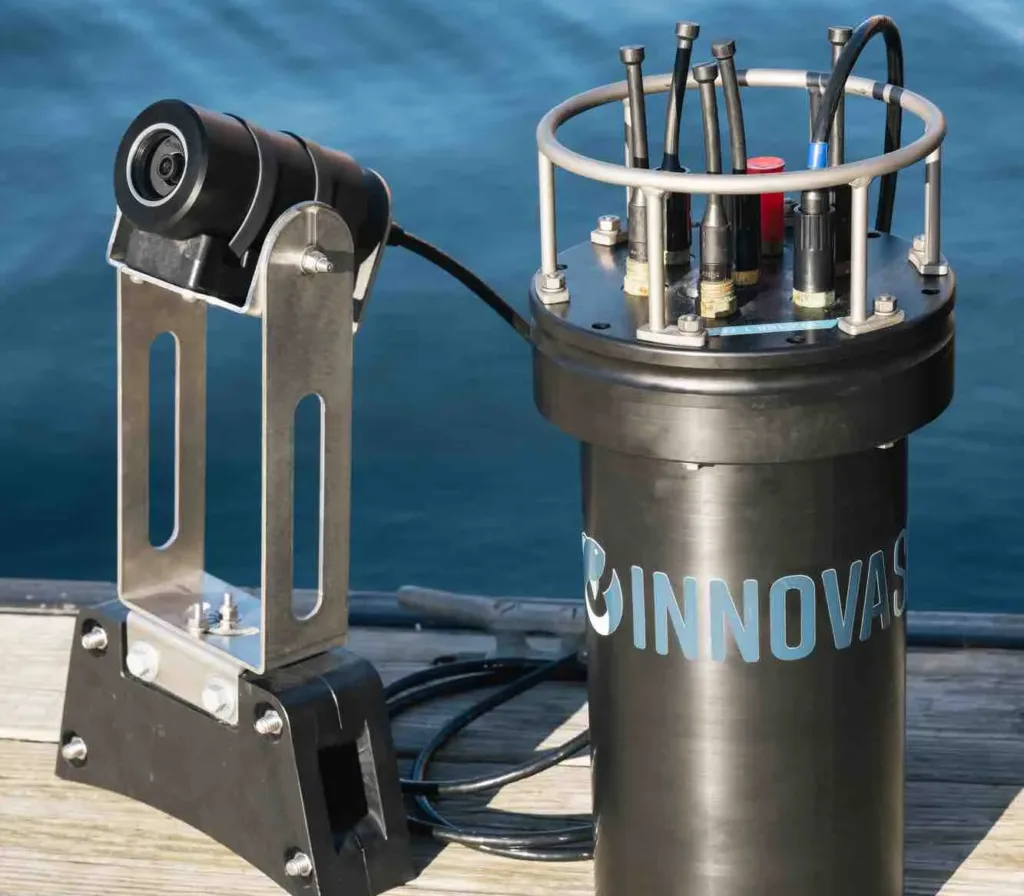 Scientific water sampling equipment on wooden dock, featuring Innovasea branded cylindrical device with sensors and mounting bracket over blue water.