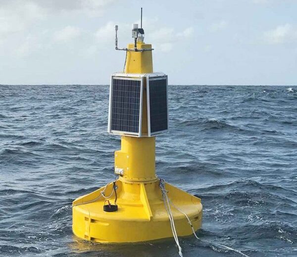 A yellow ocean buoy with solar panels and antenna equipment floating on choppy blue water under a cloudy sky.