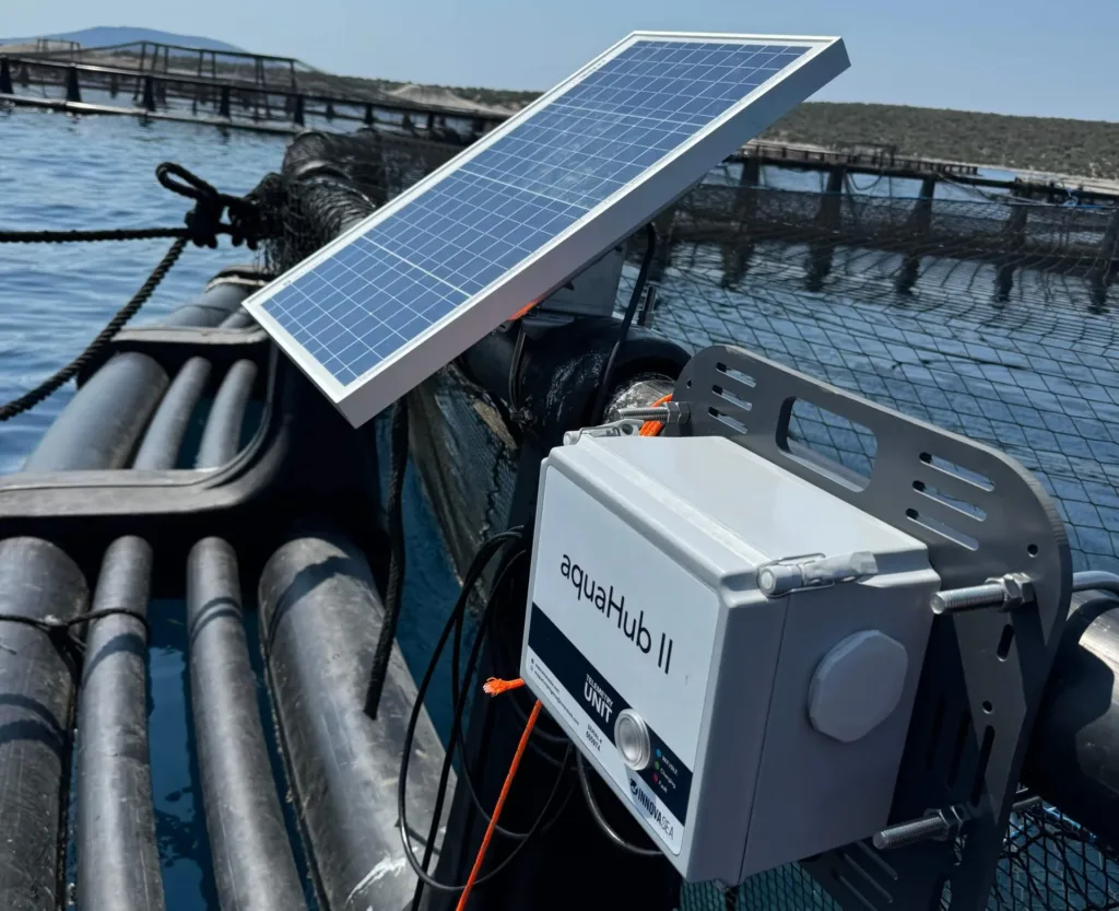 Solar-powered aquaculture monitoring device labeled "aquaHub II" mounted on fish farm cages in coastal waters with nets and equipment visible.