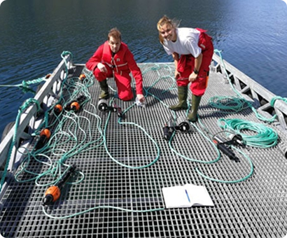 Two researchers in red gear examine scientific equipment on a boat deck over dark water.
