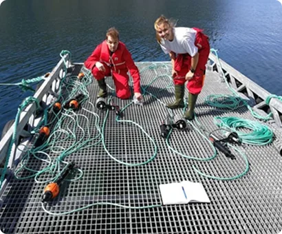 Two researchers in red gear examine scientific equipment on a boat deck over dark water.