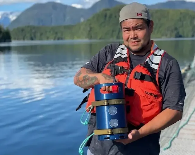 Man in life jacket holds blue cylindrical scientific equipment on boat deck with mountains and lake in background.