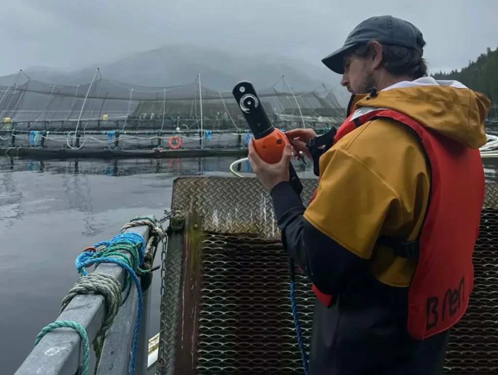 A man on a dock with an aquaMeasure sensor in yellow and red safety gear operates handheld equipment on a boat near fish farming pens in misty water.