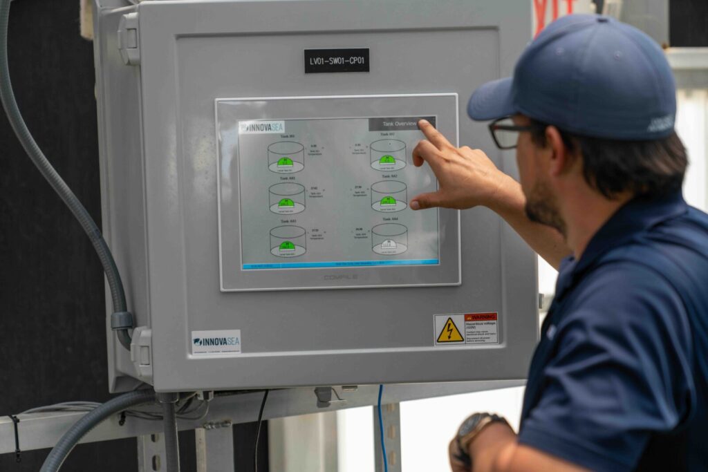 Worker in blue uniform and cap operates touch screen control panel showing tank overview with green status indicators on industrial equipment.