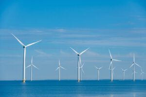 Offshore wind farm with multiple white wind turbines standing in blue ocean water under a clear blue sky.