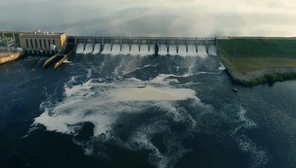 Large concrete dam with multiple spillways releasing water into turbulent river below, creating white foam and rapids.