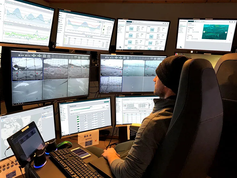 Person monitoring multiple computer screens displaying charts, data, and underwater camera feeds from a control room chair.