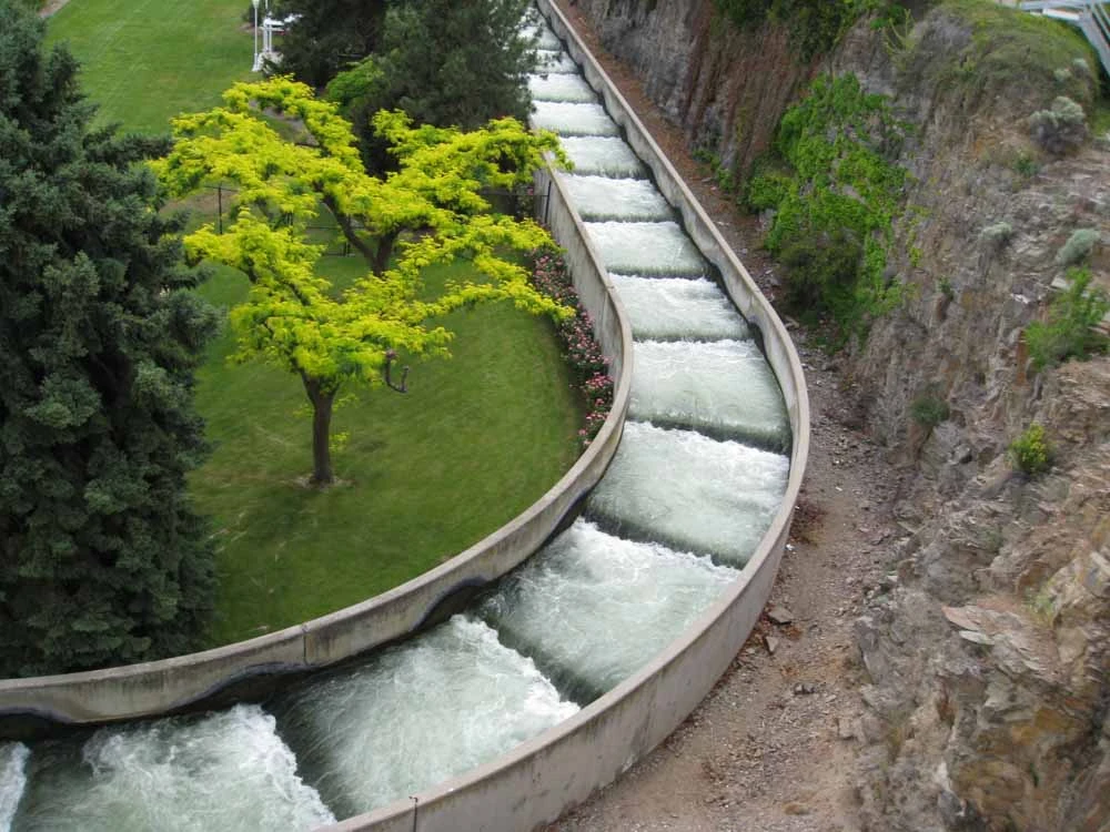 Curved concrete water channel with stepped spillways flowing through a landscaped area with green lawn and trees.