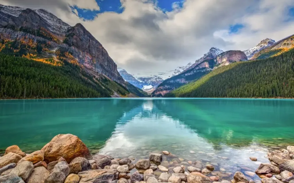 Turquoise mountain lake with rocky shore, surrounded by snow-capped peaks and evergreen forests under dramatic cloudy sky.