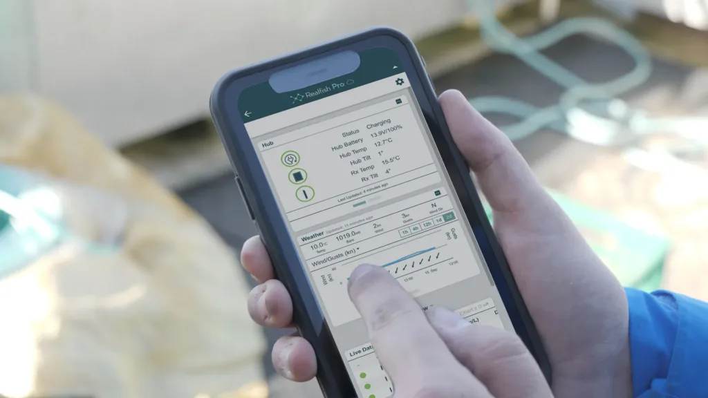 Person holding smartphone displaying Realfish Pro app with weather data, hub status, battery level, and wind/temperature charts on screen.