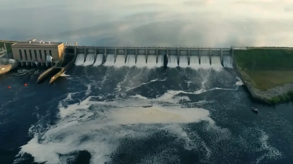 Large concrete dam with multiple spillway gates releasing water into a turbulent river below, creating white foam and spray.