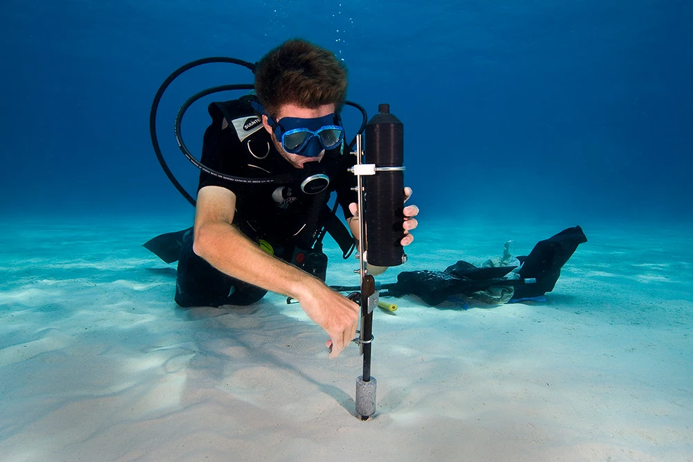 Scuba diver in full gear using a core sampling tool to extract sediment from the sandy ocean floor underwater.
