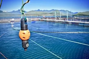 A marine sensor device suspended by rope over blue ocean water near aquaculture fish farming pens with mountains in the background.
