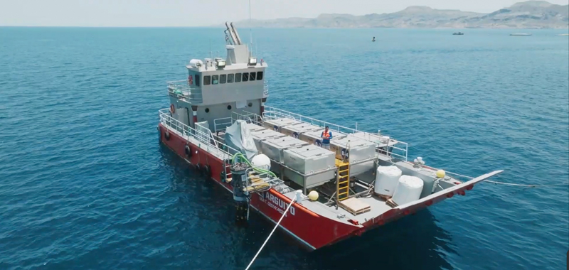 Red and white research vessel with equipment containers anchored in blue ocean waters near mountainous coastline.