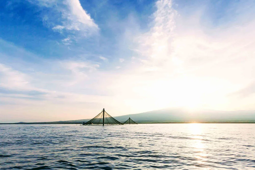 Seastation at sunset spans across calm water under a cloudy blue sky with sunlight breaking through on the horizon.