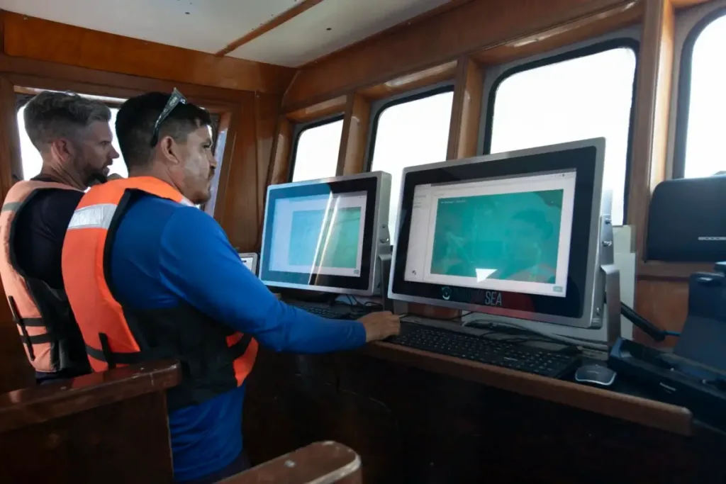 Two crew members in life jackets operate sonar equipment on multiple computer screens aboard a research vessel.