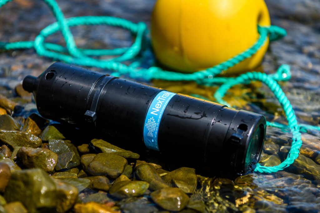 Black waterproof flashlight with blue Nexfra label lying on wet rocks near a yellow buoy and green rope.