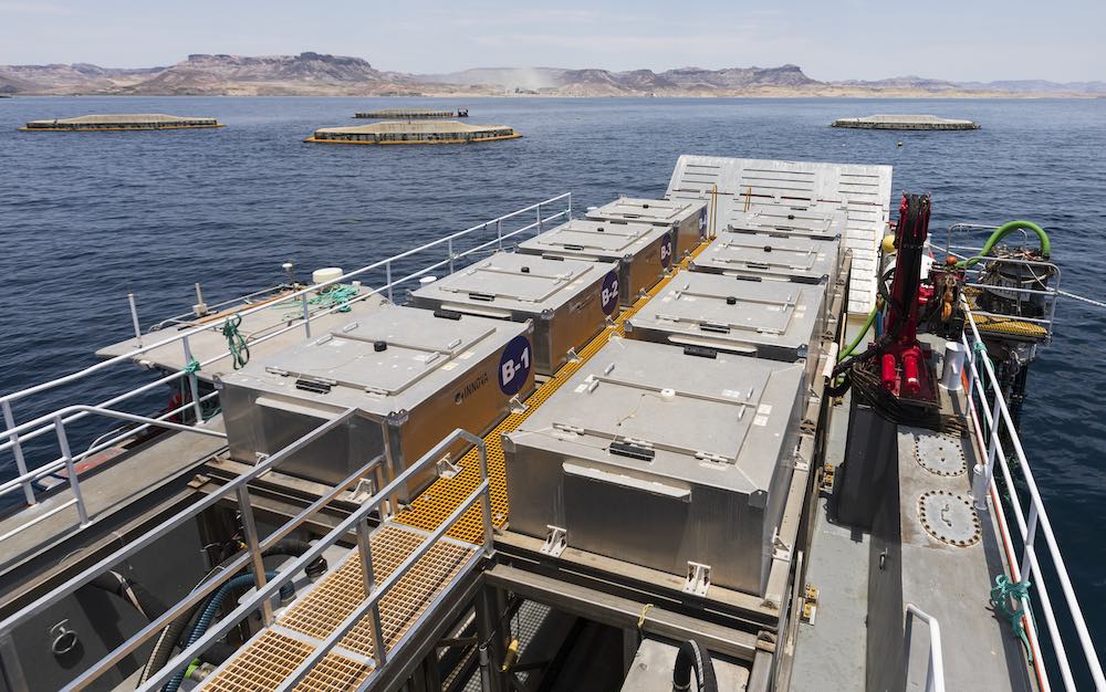 A research vessel deck with multiple white sampling containers and equipment, surrounded by water with fish farming pens visible in the background near a mountainous coastline.