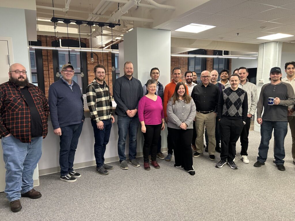 Large group of approximately 20 office employees posing together in a modern workplace with exposed brick walls and industrial lighting.