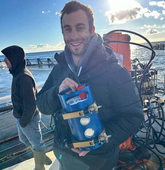 A smiling man in a dark jacket holds blue scientific equipment while standing on the deck of a research vessel at sea.